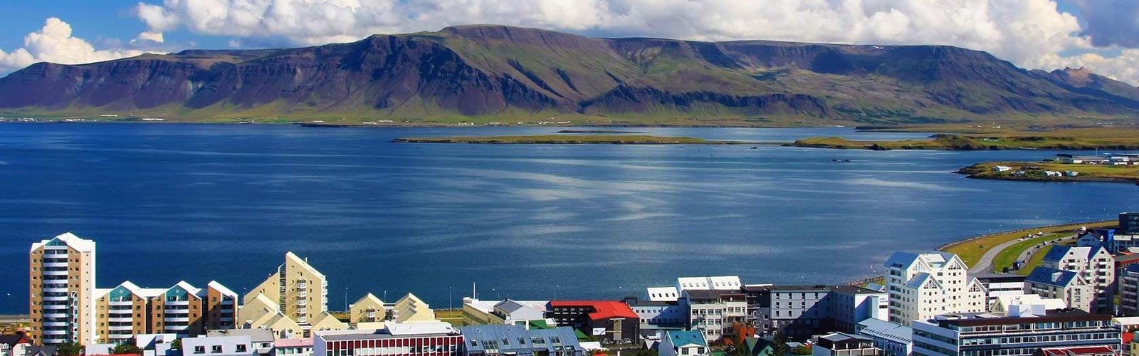 A green mountain behind a dark blue lake. The colorful buildings and rooftops of Reykjavik are just peaking out in the foreground. 